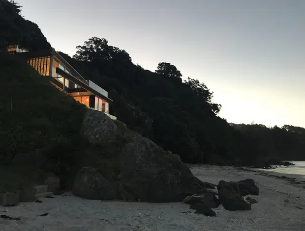 Dusk exterior view of a contemporary multi-level house built into a steep, heavily vegetated coastal hillside. The building has long horizontal glazing, warm interior lighting, dark structural framing, and light wall panels, with large exposed rocks and a sandy, rocky beach in the foreground.