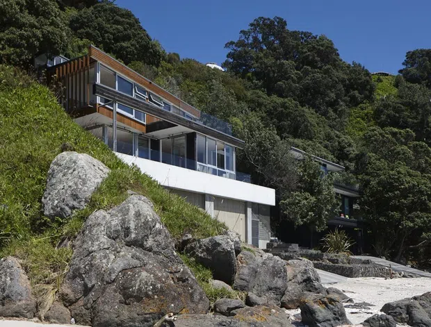 Exterior view of a contemporary multi-level hillside house built into a steep, rocky, heavily vegetated site. The home features extensive glazing, projecting balconies and decks, horizontal roof planes, timber-toned vertical cladding, and exposed concrete elements. Large boulders and a rocky shoreline or landscape edge occupy the foreground.