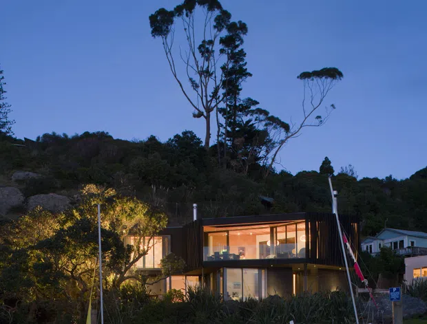 Twilight exterior view of a contemporary two-storey home set against a wooded hillside. The house has dark vertical cladding, extensive warm-lit floor-to-ceiling glazing, and a partially projecting upper level. A wet waterfront edge or beach-like foreground with small sailboats and a slipway is visible, along with neighbouring homes and native trees.