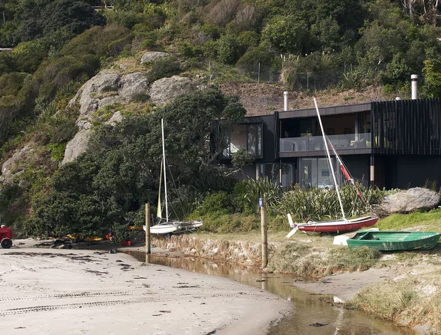 Exterior view of a dark, modern multi-level house set into a rocky, densely vegetated coastal or waterfront site. The building has a rectilinear form, extensive glazing, dark vertical cladding, balconies or projecting decks, and a low-profile roofline. Small boats and a sandy shoreline are visible in the foreground.