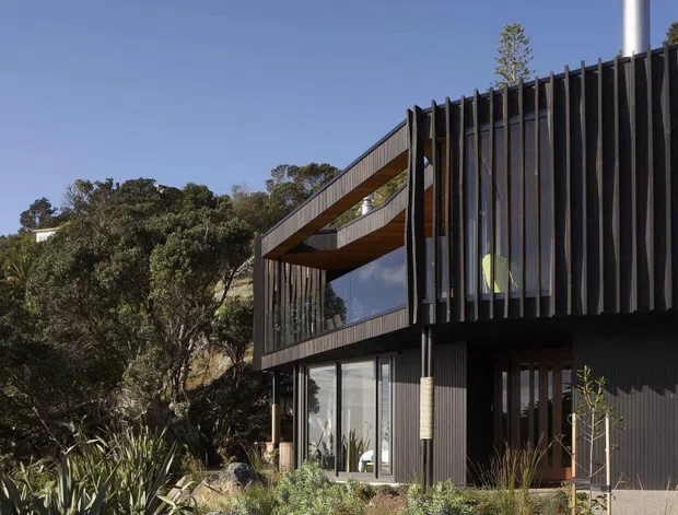 Exterior view of a contemporary two-storey house set into a natural, rocky landscaped site. The building has dark vertical cladding, expansive glazing, projecting upper-level balconies or terraces, timber soffits, and a prominent metal flue. Native-style planting, grasses, rocks, and mature trees surround the facade under a clear blue sky.