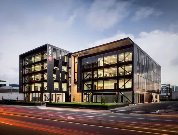 Exterior view of a contemporary multi-storey commercial office building at dusk, with two connected glass-fronted volumes, dark metal framing and cladding, warm interior lighting, and prominent street-level traffic light trails in the foreground.