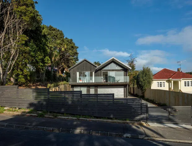 Street-facing exterior view of a contemporary two-storey home with twin gabled roof forms, dark vertical cladding, extensive upper-level glazing and balconies, and a dark horizontal-slat boundary fence and gate. A neighbouring traditional house and mature trees are visible alongside the property.