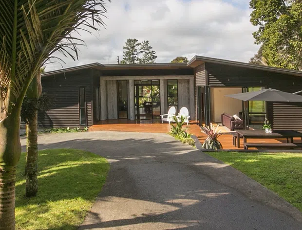Single-storey contemporary home exterior set in a landscaped tropical garden. The dark horizontal-clad facade has broad glazing, a recessed central entry, low-pitched roof forms, and a timber entertaining deck with outdoor seating and umbrella. A paved driveway or walkway leads through the lawn toward the house.