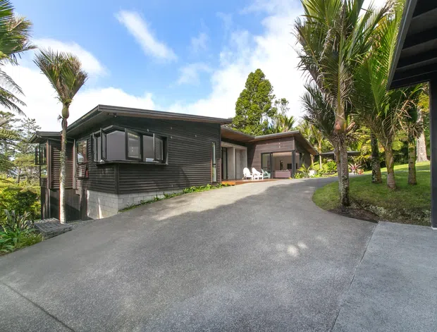 Exterior view of a low-rise dark-clad house set among lush tropical planting. The building has a predominantly single-storey, low-pitched roof form, horizontal dark wall cladding, projecting window bays, and a broad concrete driveway rising toward the entrance and covered area. Palm trees and landscaped lawns surround the home, with a bright blue sky and sunny conditions.