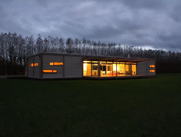 A low, elongated modern single-storey dwelling set in an open grassy landscape at dusk. The building has dark horizontal cladding, a shallow flat or low-pitched roof, extensive warm-lit glazing, and a covered recessed central outdoor living area framed by slender posts. Tall trees form a wooded backdrop beneath a dramatic overcast sky.