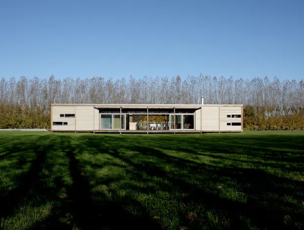 A low, elongated modern single-storey residence set within a broad grassy landscape. The facade combines pale horizontal cladding with large central glazing and projecting roof planes, creating a strong indoor-outdoor connection. Deciduous trees form a backdrop, while long shadows fall across the lawn under a clear blue sky.