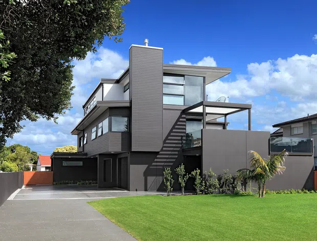 Exterior view of a contemporary multi-storey residence with dark horizontal cladding, extensive glazing, projecting flat roof forms, an external stair, and a broad lawn and driveway in front. The strongly geometric facade is set within a suburban landscape under a bright sky.