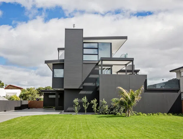 Exterior view of a contemporary multi-level residence with strong geometric forms, dark grey cladding, expansive glazing, projecting flat roof planes, and layered balconies or terraces. The house is set behind a manicured lawn with low planting and neighbouring suburban homes visible around it.