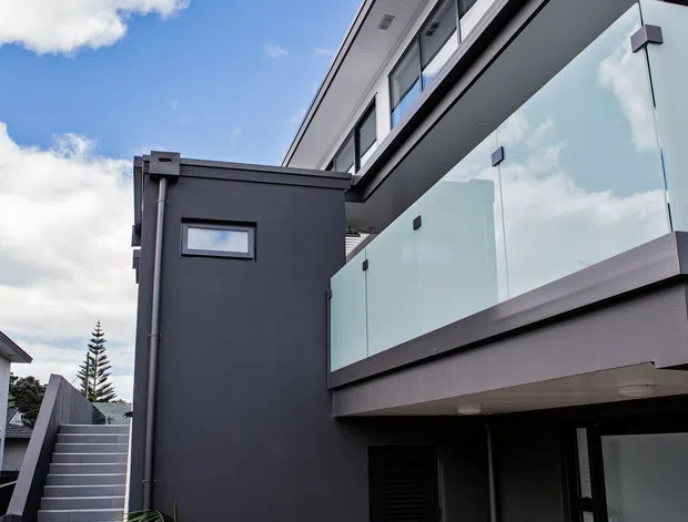 Exterior view of a contemporary multi-level home with dark charcoal cladding, projecting flat rooflines, extensive glazing, and a frosted glass balcony balustrade. An external stair leads to the upper level, while a timber deck, tropical planting, and outdoor seating occupy the courtyard-like foreground.