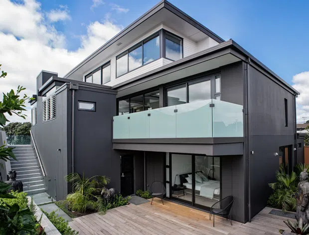 Contemporary multi-level residential home with a dark charcoal rendered facade, flat rooflines, extensive glazing, and a projecting upper-level balcony with frosted glass balustrade. A timber-toned deck and landscaped garden surround the lower living area, while an external stair leads to the upper level.