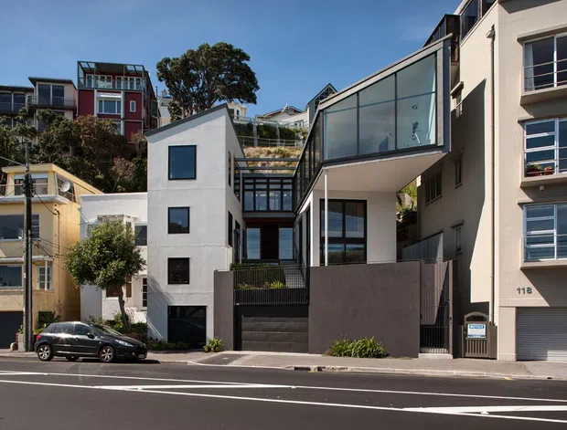 Street-level view of a contemporary multi-storey urban residence on a sloping site, featuring crisp white rendered volumes, extensive dark-framed glazing, a projecting glazed upper floor, dark privacy fencing, and stepped landscaping. Adjacent apartment and residential buildings line the street, with overhead utility wires and a marked roadway in the foreground.