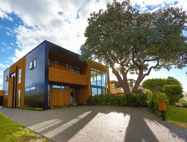 Exterior view of a contemporary multi-level residential house with dark vertical cladding, warm timber wall panels, extensive glazing, and a flat roof. The home fronts a paved driveway and landscaped verge, with a mature street tree and suburban road visible to the right.