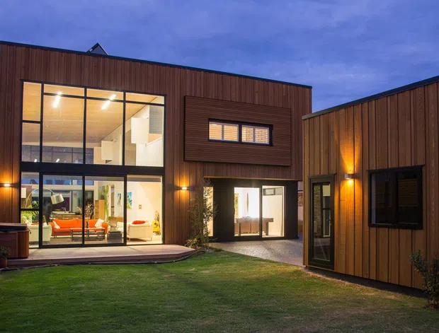 Twilight exterior view of a contemporary courtyard-style residence with warm vertical timber cladding, flat roof forms, extensive floor-to-ceiling glazing, and illuminated interior spaces opening onto a small lawn. The composition highlights the angular multi-wing building and sheltered courtyard.