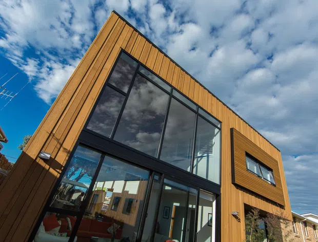 Low-angle exterior view of a contemporary multi-storey residence clad in warm vertical timber boards. A large angular section of black-framed glazing forms the main facade, with additional projecting window surrounds and a glazed entry or living area visible at ground level. The sky and neighbouring buildings surround the strong geometric facade.