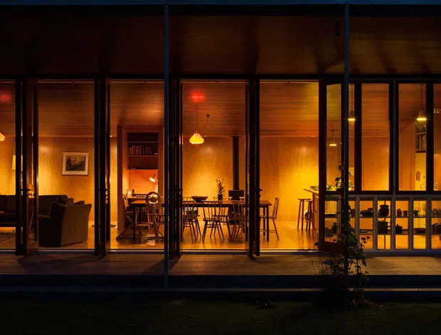 Night-time exterior view through full-height glazing of a warmly illuminated modern home. The composition shows a timber-lined interior with a living area, dining area, pendant lights, dark-framed glazing, and a timber deck, creating a strong indoor–outdoor connection.