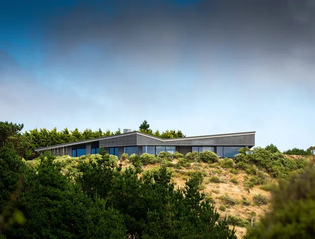 Low-profile modern single-storey building set into a densely vegetated, sandy coastal-looking landscape. The long horizontal form has extensive dark-framed glazing, a shallow pitched or folded roofline, and dark grey exterior cladding. Shrubs and dune-like planting dominate the foreground, with an expansive cloudy sky above.