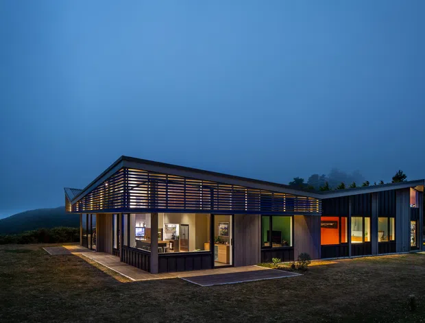 A low, modern single-storey house photographed at dusk, set against a dark wooded and mountainous backdrop. The building has a crisp rectilinear form with dark vertical cladding, extensive glazing, and a prominent projecting roof or canopy wrapped in horizontal timber-like screening. Warm interior lighting glows through the windows and highlights the sheltered entry and patio areas.