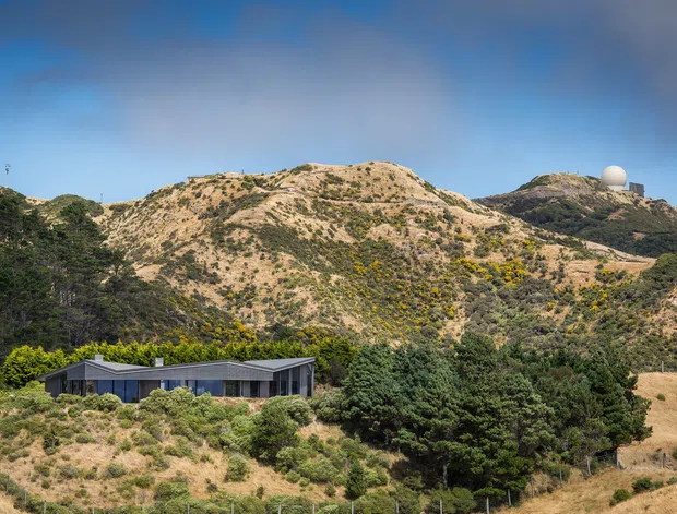 A low, modern building sits in a dry, rugged hillside landscape beneath a bright blue sky. The structure has a dark horizontal roofline, extensive glazing, and a muted grey facade, surrounded by native-looking vegetation and rolling scrub-covered hills. A spherical structure is visible on a distant ridgeline.