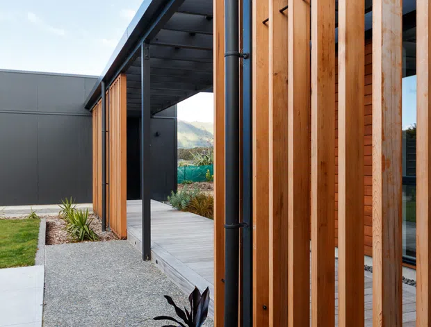 Exterior architectural view of a contemporary building entry or covered walkway. The facade combines vertical timber screening with dark metal framing and a flat roof, alongside gravel landscaping, planted garden beds, and concrete and timber-paved paths. The open passage leads toward a landscaped courtyard, with distant hills visible beyond.