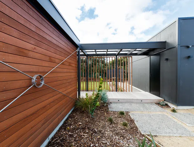 Exterior view of a contemporary residential entry or side passage. A warm horizontal timber-clad wall with a distinctive tension-cable detail contrasts with dark grey metal-clad volumes. A glazed or translucent-roofed covered walkway leads toward a landscaped courtyard, with vertical timber screening, concrete stepping slabs, gravel, and garden beds.