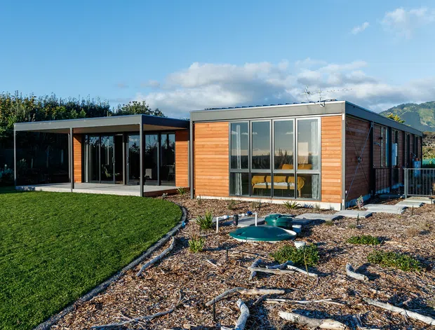 Exterior view of a low, single-storey contemporary home set within a landscaped garden. The building combines warm horizontal timber-clad walls with extensive dark-framed glazing, a projecting flat-roofed glazed living pavilion, and a simple flat-roofed main volume. Natural mulch planting, lawn, and sculptural driftwood landscaping occupy the foreground, with a wooded hillside visible in the distance.