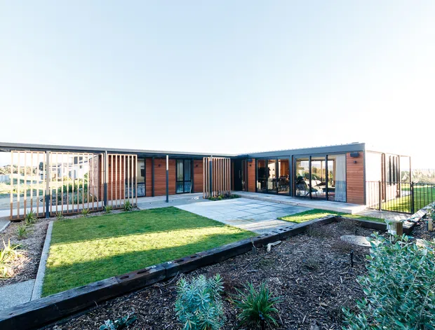 Single-storey contemporary courtyard-style residence with a low, mostly flat roofline, warm timber-clad wall sections, extensive black-framed glazing, and vertical timber privacy screens. The home wraps around a paved central courtyard and lawn, with landscaped garden beds in the foreground and a covered outdoor area visible through the glazing.