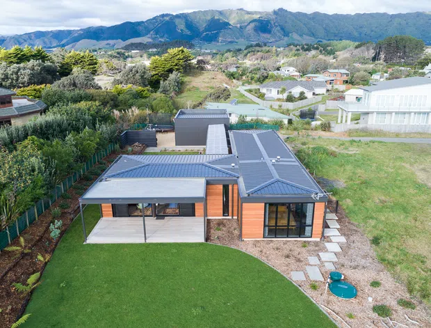 Aerial view of a contemporary single-storey house with a dark pitched metal roof, warm timber-toned cladding, and extensive glazing. The home is set within landscaped gardens with a lawn, planted borders, paved paths, and a small plunge pool. A suburban/rural residential setting and mountain range are visible in the background.