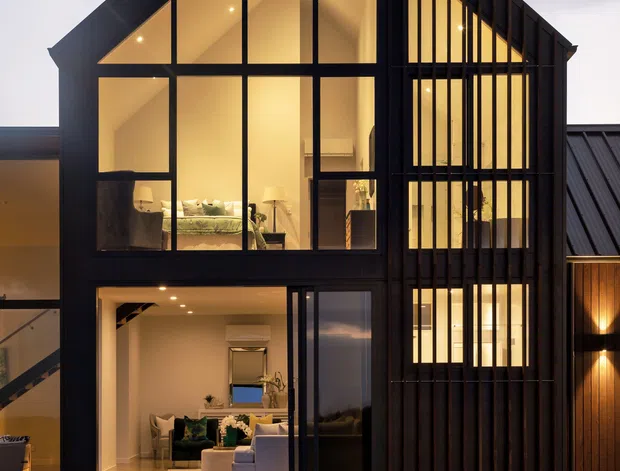 Twilight exterior view of a contemporary multi-storey house with a steep gabled roof and dramatic black-framed floor-to-ceiling glazing. Warm interior lighting reveals living spaces through the windows, while vertical dark cladding and fins create a strong linear facade.