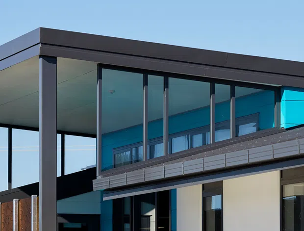 Exterior architectural view of a contemporary multi-level residence with a flat roof, strong horizontal lines, dark metal framing, expansive floor-to-ceiling glazing, and light wall cladding. A covered terrace or carport-like structure projects from the main volume, with vivid blue-tinted glazing visible along the upper level.