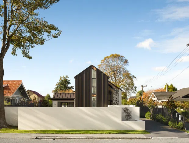 Street-facing view of a contemporary multi-level residential house with a tall, narrow dark-clad gabled volume, vertical glazing, and a low sculptural white rendered boundary wall. The home sits within an established suburban neighbourhood alongside older pitched-roof houses, mature trees, a driveway, and overhead utility lines.