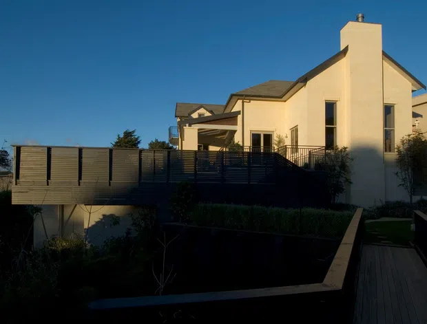 Exterior view of a contemporary light-rendered house at dusk or in low evening light, featuring a pitched roof, prominent chimney, projecting timber deck, horizontal privacy screens, dark metal railings, and a landscaped sloped garden with a timber walkway.