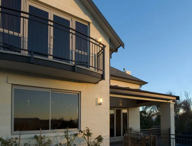 Exterior view of a two-storey contemporary residence with pale masonry cladding, dark rooflines, large glazed openings, an upper-level balcony with black metal balustrade, and a covered entry terrace. Landscaping and a glimpse of a pool or dark outdoor area appear at the lower right.