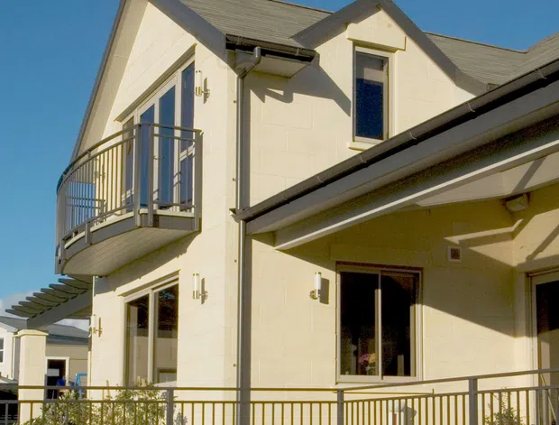 Exterior view of a light-coloured multi-storey residential building with a pitched gabled roof, projecting balcony, glazed doors and windows, covered terrace, and metal railings. The facade has cream painted masonry or rendered blockwork with grey roofing and trim, photographed in bright daylight.