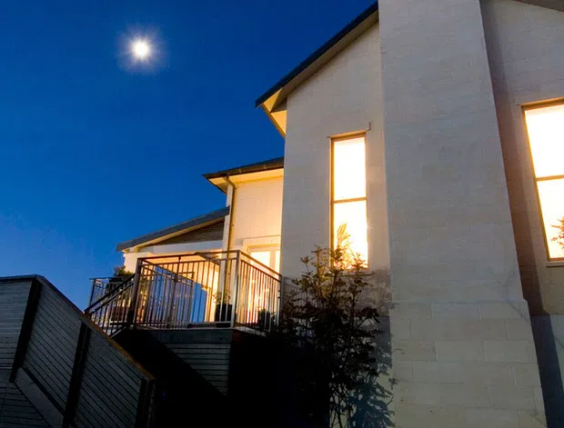 Twilight exterior view of a contemporary multi-level home with pale masonry walls, dark pitched rooflines, a tall chimney-like vertical feature, illuminated windows, and a raised balcony with metal railing. The home is set on a sloping site, with neighboring roof forms and landscaping visible in the foreground.