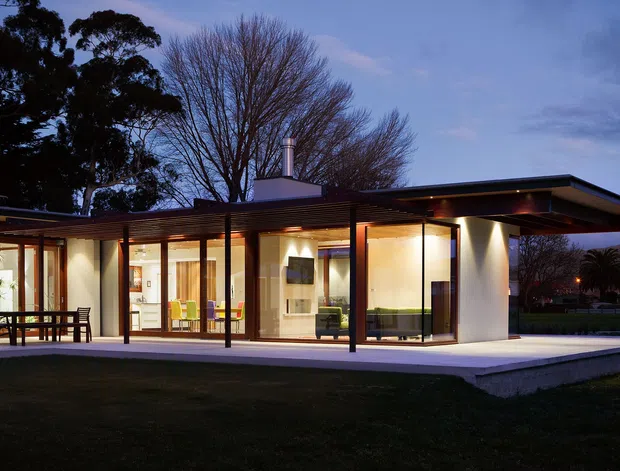 Single-storey modernist home photographed at dusk, with a low flat roof, broad cantilevered eaves, extensive floor-to-ceiling glazing, slender dark structural posts, and a covered terrace wrapping the living spaces. Warm interior lighting reveals an open-plan living and dining area, while mature trees and lawn surround the house.