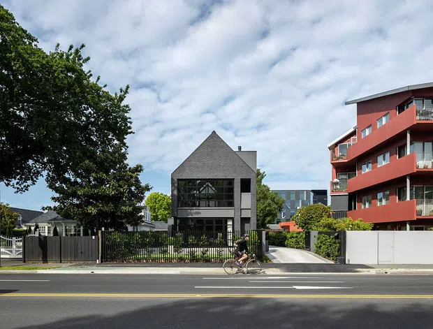 Street-level view of a contemporary urban residential development, with a dark grey gabled townhouse or house at centre and a multi-storey red apartment building with projecting balconies to the right. Front fences, landscaped gardens, a driveway, footpath and road occupy the foreground, with mature trees and additional buildings in the background under a partly cloudy sky.