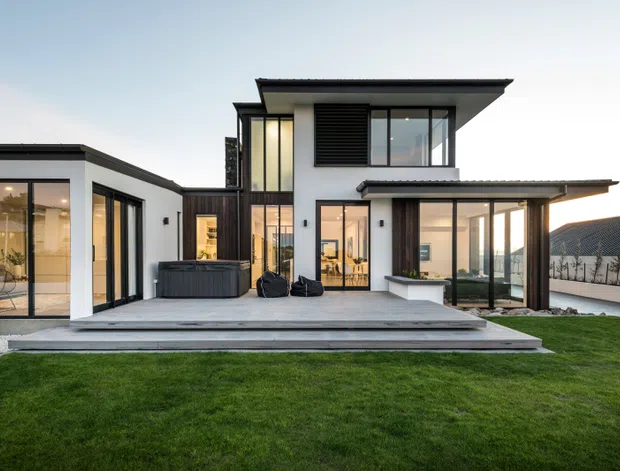 Exterior view of a contemporary two-storey residence with crisp white rendered walls, dark metal roof planes, extensive floor-to-ceiling glazing, timber-toned vertical cladding, and a broad timber or composite deck opening onto a manicured lawn. The composition includes a projecting covered outdoor living area and a low single-storey glazed wing.