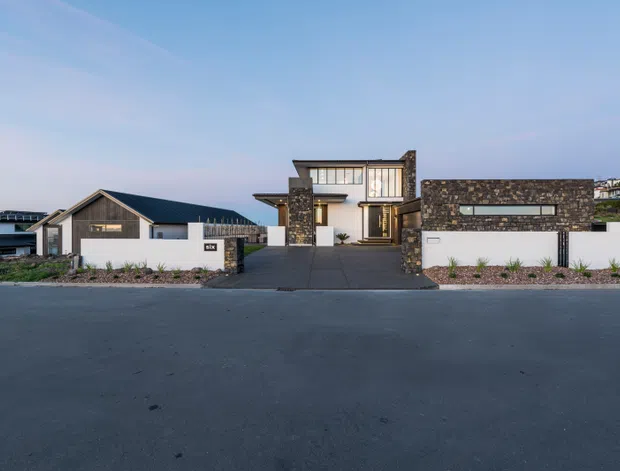 Street-facing view of a contemporary multi-level residence with dark stone cladding, white rendered walls, large glazed windows, a dark pitched roof, and a broad paved driveway. A separate gabled structure is visible to the left, with low landscaped planting along the frontage.