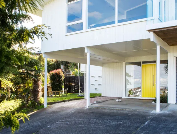 Exterior view of a raised two-storey residential house with a white vertically ribbed facade, broad floor-to-ceiling glazing, a bright yellow entry door, and a covered open carport or driveway area beneath the upper level. A small balcony with clear glass balustrading is visible at right, with landscaped planting around the paved frontage.