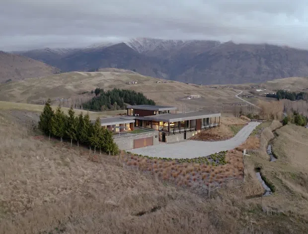 Aerial view of a contemporary low-rise residence set within a broad, rugged mountain landscape. The home has a long horizontal composition, dark low-pitched roof forms, extensive glazing, warm timber-toned exterior elements, and a curved driveway approaching the building. Native-looking landscaped planting surrounds the house, with dramatic barren mountains and a cloudy sky forming the backdrop.