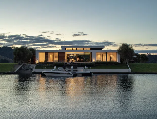 Wide exterior view of a low, contemporary single-storey waterfront residence at dusk. The house has a long rectilinear form, extensive floor-to-ceiling glazing, a shallow flat roof with a raised clerestory-like central section, warm interior lighting, landscaped lawn, and a private dock with a small boat along the water.