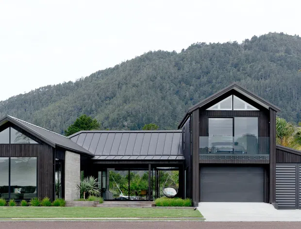 Exterior view of a contemporary multi-volume residence with dark vertical cladding, standing-seam metal gabled roofs, large glazed openings, a prominent glazed upper-level gable, and an integrated garage. The home sits against a wooded mountain backdrop with palms and landscaped lawn.