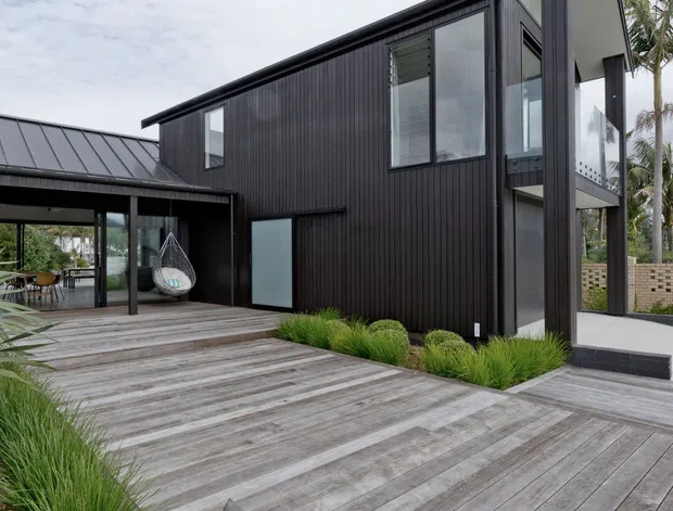 Exterior view of a contemporary two-storey dark-clad home with a sharply angled roofline, extensive glazing, and a broad weathered timber deck. A covered outdoor entertaining area connects to the main building, with landscaped grasses and glimpses of palm vegetation around the site.