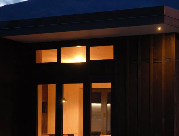 Exterior dusk view of a compact dark-clad modern building with a broad flat roof and warm interior lighting visible through tall windows and upper clerestory openings. A dramatic mountain silhouette and cloudy sky form the background. The facade appears vertically clad in dark timber or similar material, with a recessed entry and dining area visible inside.