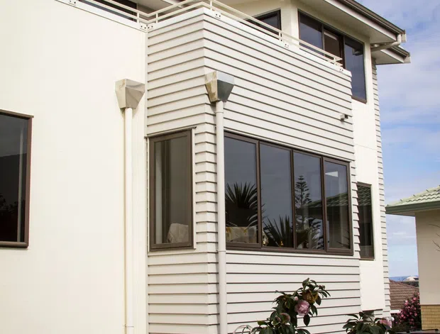 Exterior view of a multi-level residential building featuring cream horizontal weatherboard cladding, smooth light-rendered walls, dark-framed windows, and a rooftop balcony with white metal railings. Landscaped planting borders the narrow garden courtyard, with a neighbouring tiled-roof structure visible at the side.