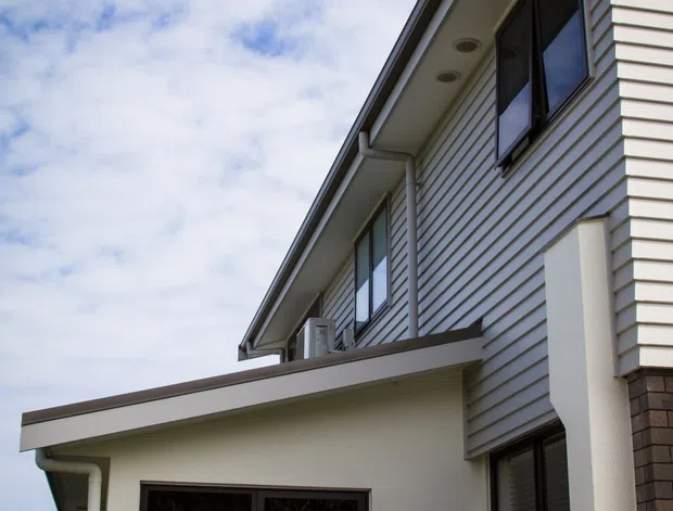 Upward-looking exterior view of a contemporary two-storey residential building. The facade combines light horizontal weatherboard cladding with smooth white rendered walls, dark-framed windows, projecting flat roof sections, exposed eaves, and a white metal balcony balustrade. A small air-conditioning unit and recessed eave lights are visible. The image is set against a partly cloudy sky.