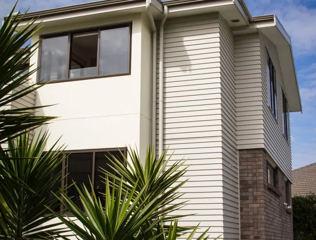 Exterior view of a contemporary multi-storey residence with cream rendered walls, white horizontal weatherboard cladding, dark-framed windows, a low-pitched roof with projecting eaves, and a lower section of brown brickwork. Tropical-style spiky landscaping partially obscures the facade.