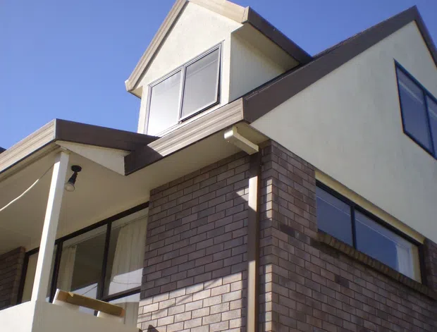 Exterior view looking upward at a multi-level residential building with dark brown brickwork, light rendered walls, projecting gabled roof forms, and large rectangular windows. A covered balcony or entry terrace with white-painted posts is visible on the left, under a clear blue sky.
