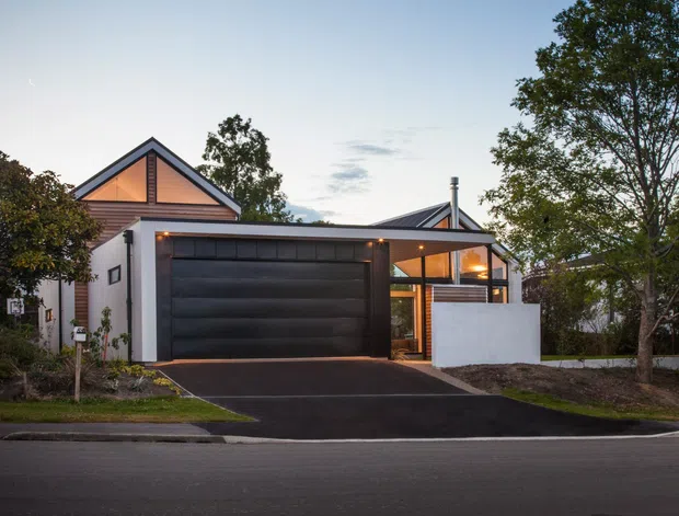 Street-facing exterior view of a contemporary residential house at dusk. The home features a prominent dark sectional garage door, a steep gabled roof with warm timber-toned cladding beneath the gable, white rendered walls, extensive glazed entry and living areas, and a small front driveway with landscaped planting and mature trees.